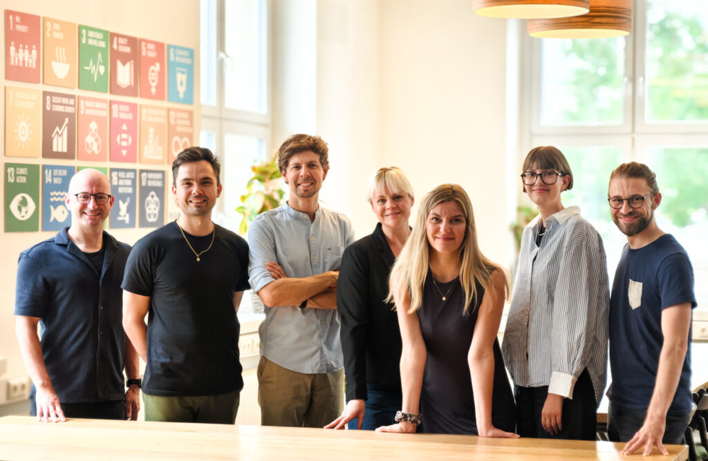 An Tisch stehend von links nach rechts die Teammitglieder Michael Jenkner, Toni Koç, Matthias Damert, Silke Nyvlt, Franziska Kramer, Emma Deeken und Steve Grundig. Sie stehen gemeinsam im Co-Working Space Impact Hub Dresden, dem Firmensitz von plant values